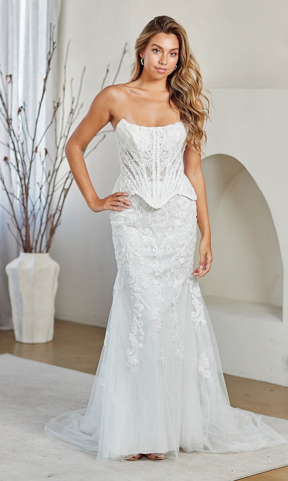 A woman stands indoors with one hand on her hip, wearing the Amelia Couture 2151 long prom dress in white, featuring a strapless design, lace embroidery, and a fitted corset bodice.